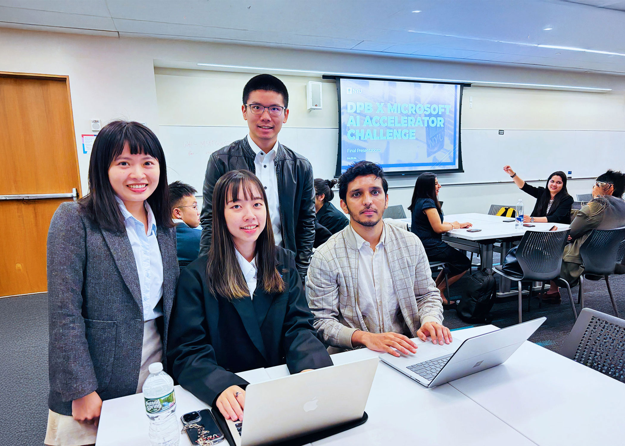 A group of four NYU SPS stidents posing at a table with laptops in a classroom setting with a presentation screen in the background for the DPB X Microsoft AI Accelerator Challenge Final Presentation.
