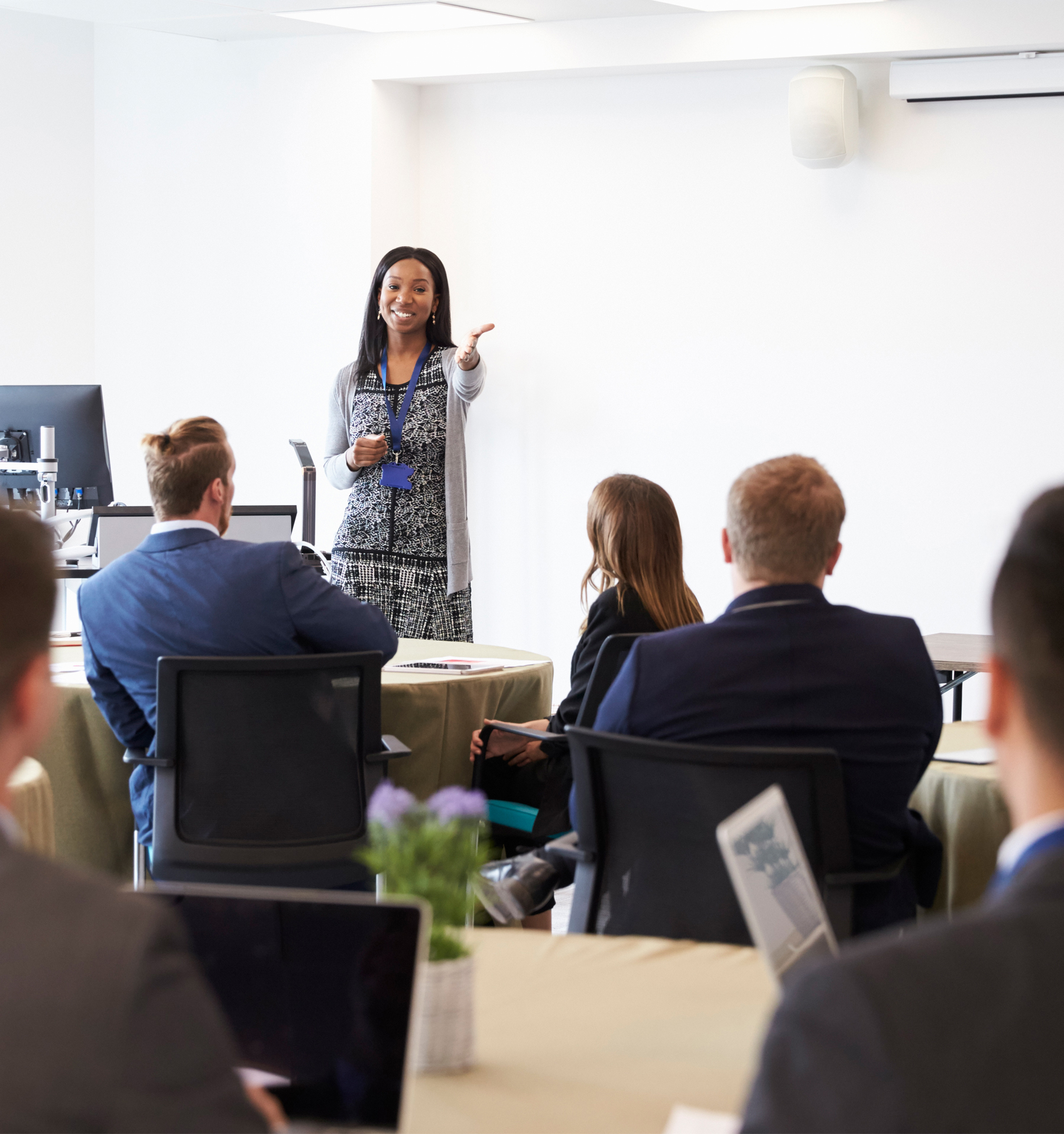 A jovial woman stands at the head of a white room, leading a presentation on organizational learning solutions in front of many engaged attendees seated at round tables.