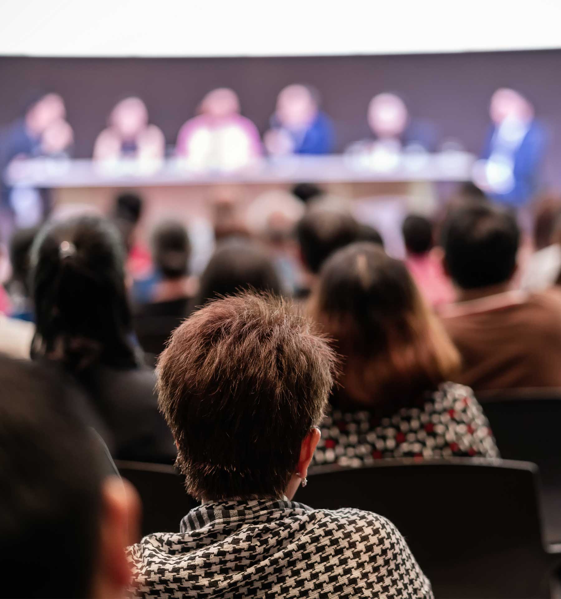 Blurred audience watching a panel of speakers at a conference. The focus is on the diverse crowd, seated and attentively engaged in a dimly lit room.