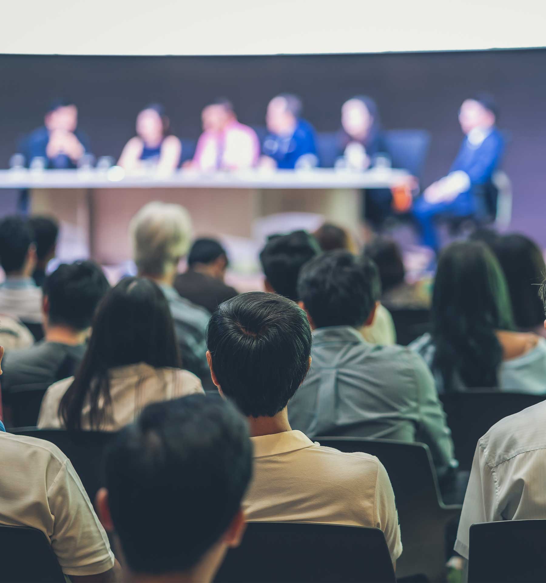 Audience listening to a panel discussion; the blurred background shows five speakers at a table on stage.