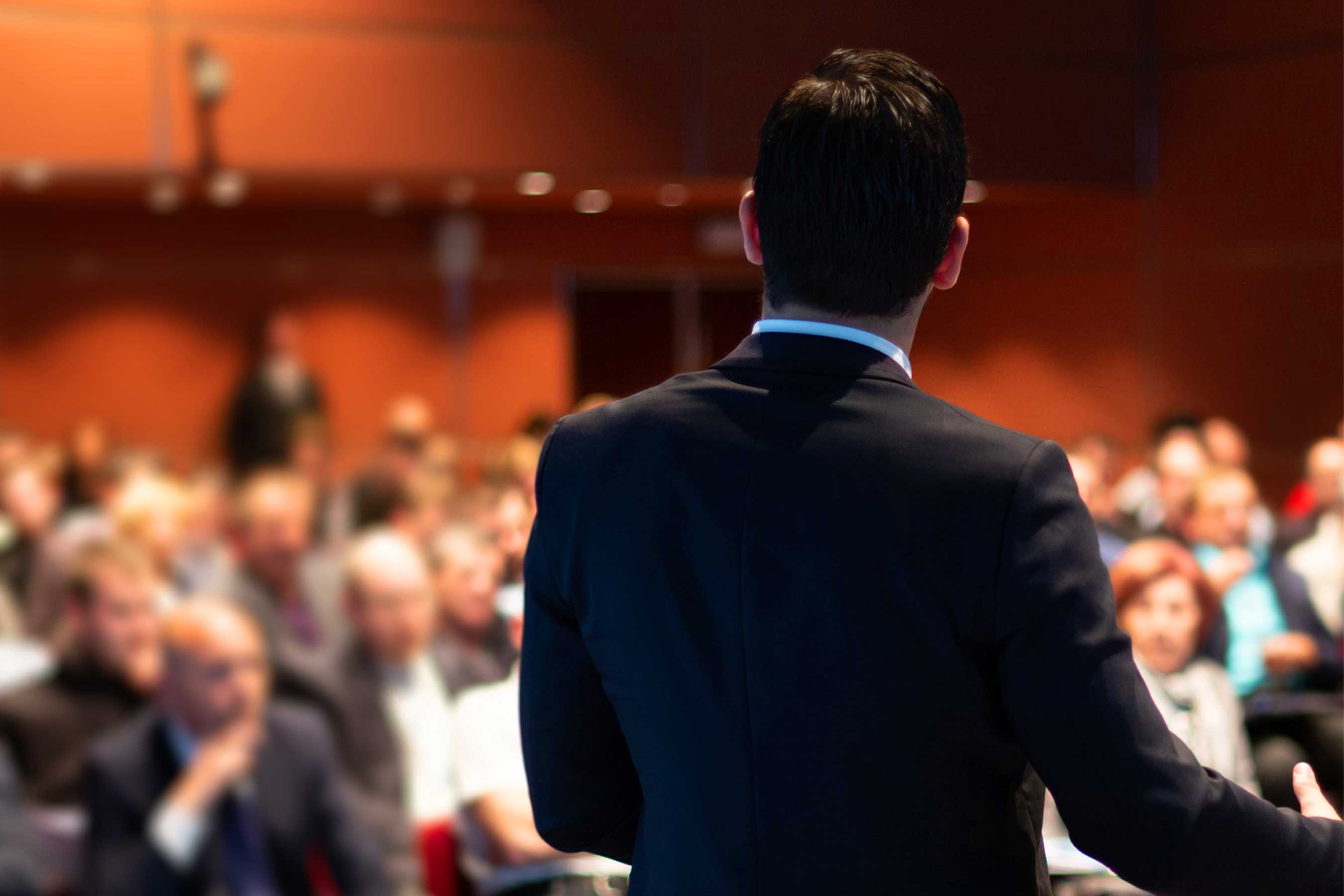 A man in a suit addresses a blurred audience in a conference room.