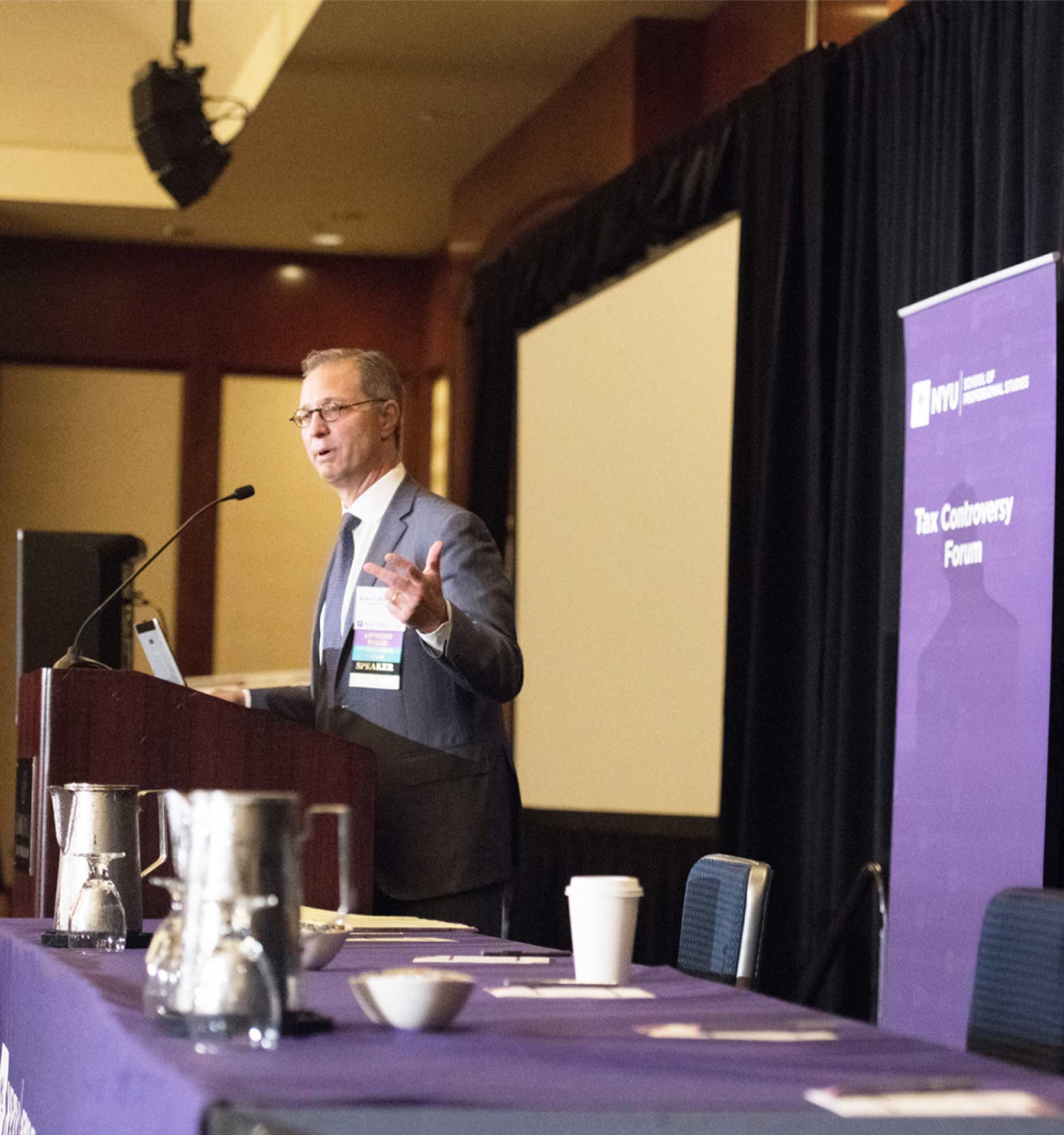 A man at a conference podium speaking and standing near a purple banner that reads "Tax Controversy Forum."
