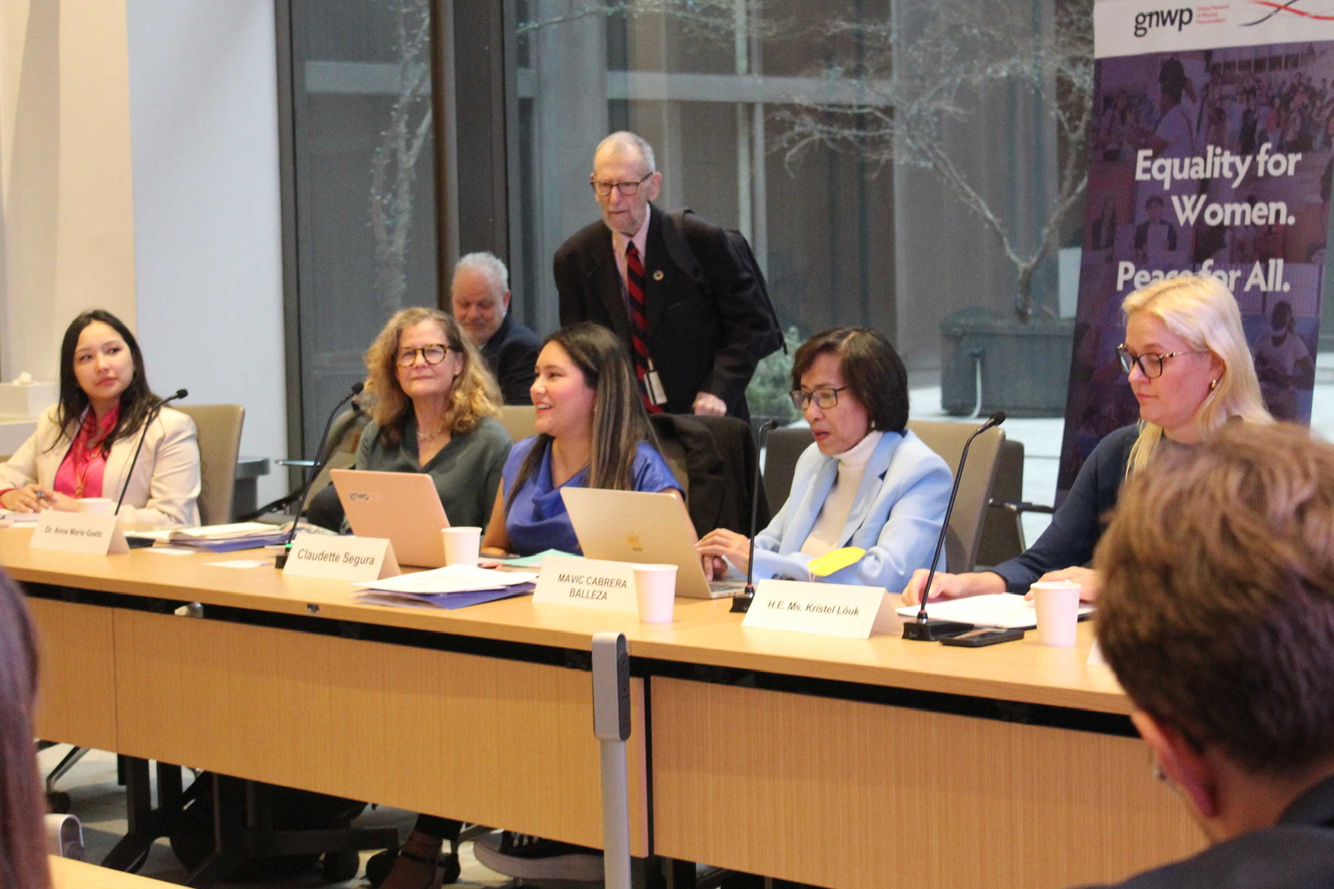 A conference room with people seated at a table, a banner reads "Equality for Women. Peace for All."