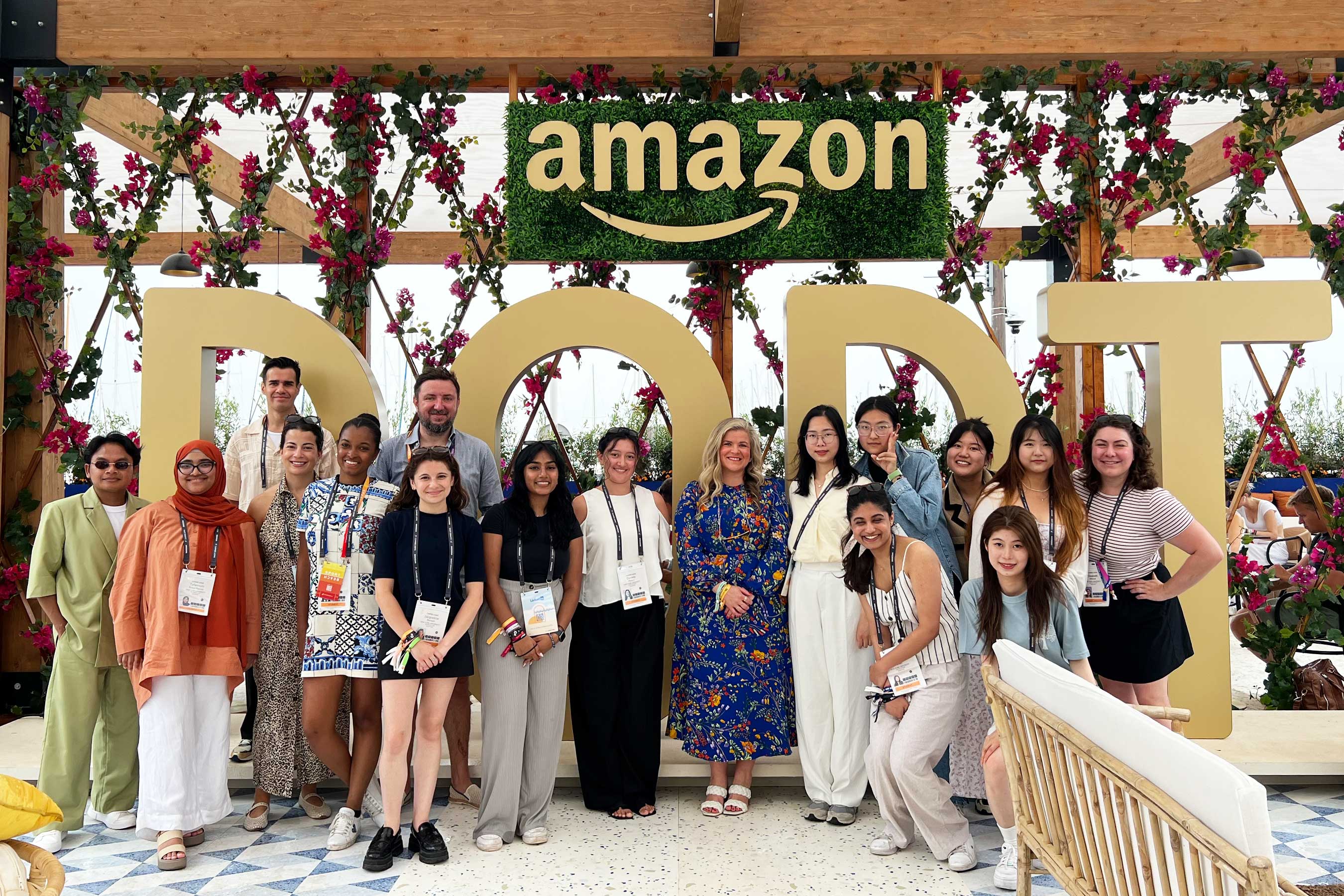 A diverse group of 16 people smiling and posing in a decorative outdoor space under a canopy, with large letters and the Amazon logo behind them.