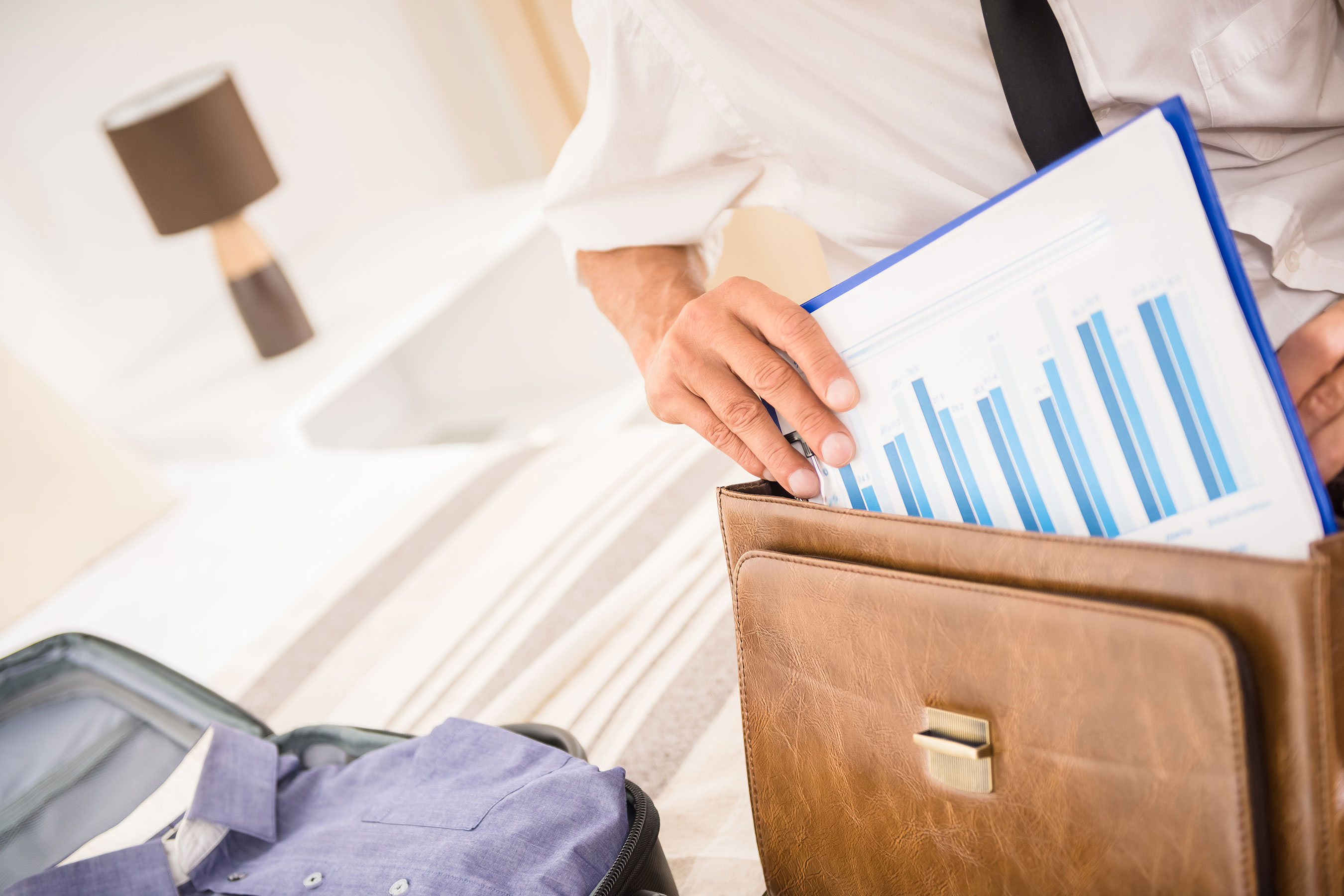 Individual pulling out statistical chart from messenger bag next to hotel bed with suitcase and packed belongings on top.