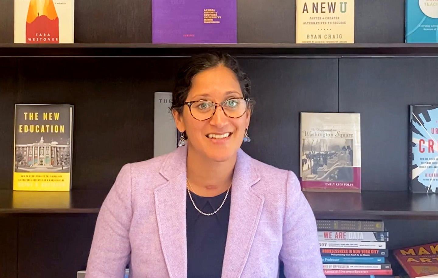 Angie Kamath, Dean of NYU School of Professional Studies, smiling while standing in front of a bookshelf filled with books.