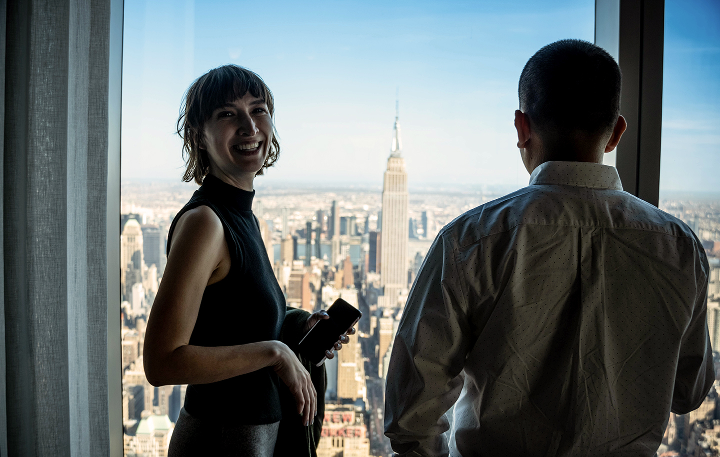 Two NYU SPS students stand in front of a large window with the Manhattan skyline in the distance while celebrating the dynamism of NYC