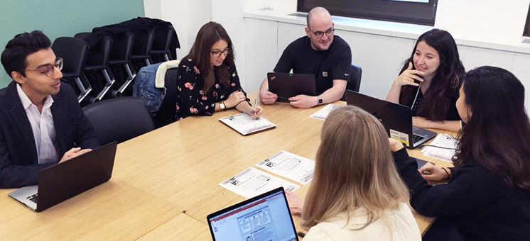 Six professionals sit around a conference table, engaged in discussion with laptops, notebooks, and printed documents.