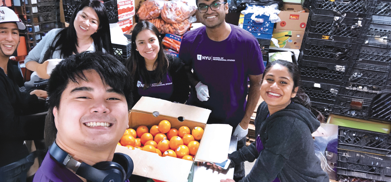 Six NYU SPS students pose for a selfie with a box of oranges at a food bank during a community service day