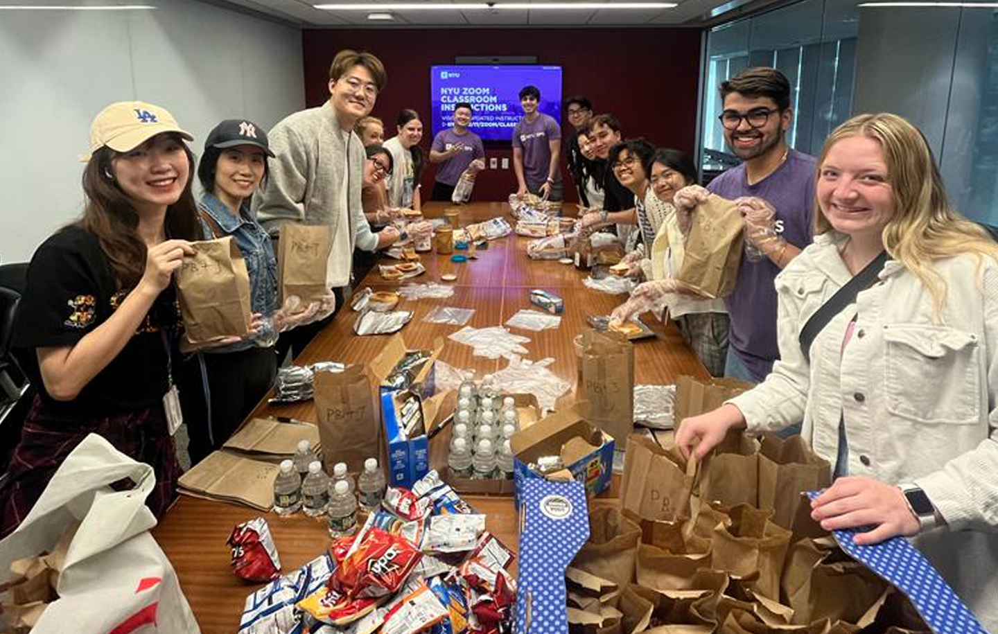 A group of NYU students pose for a photo around a long table with various food items while doing volunteer community service