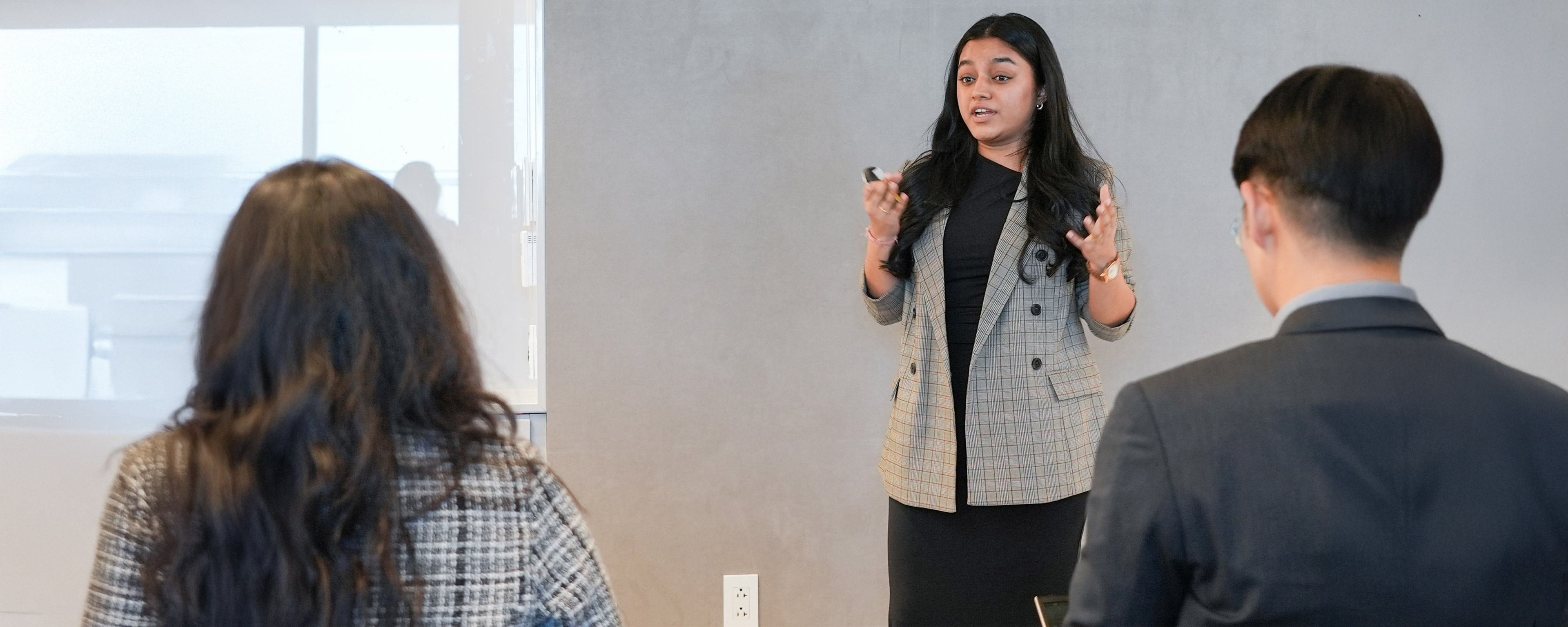 A person in a plaid blazer speaking in front of a group with a whiteboard visible in the background.