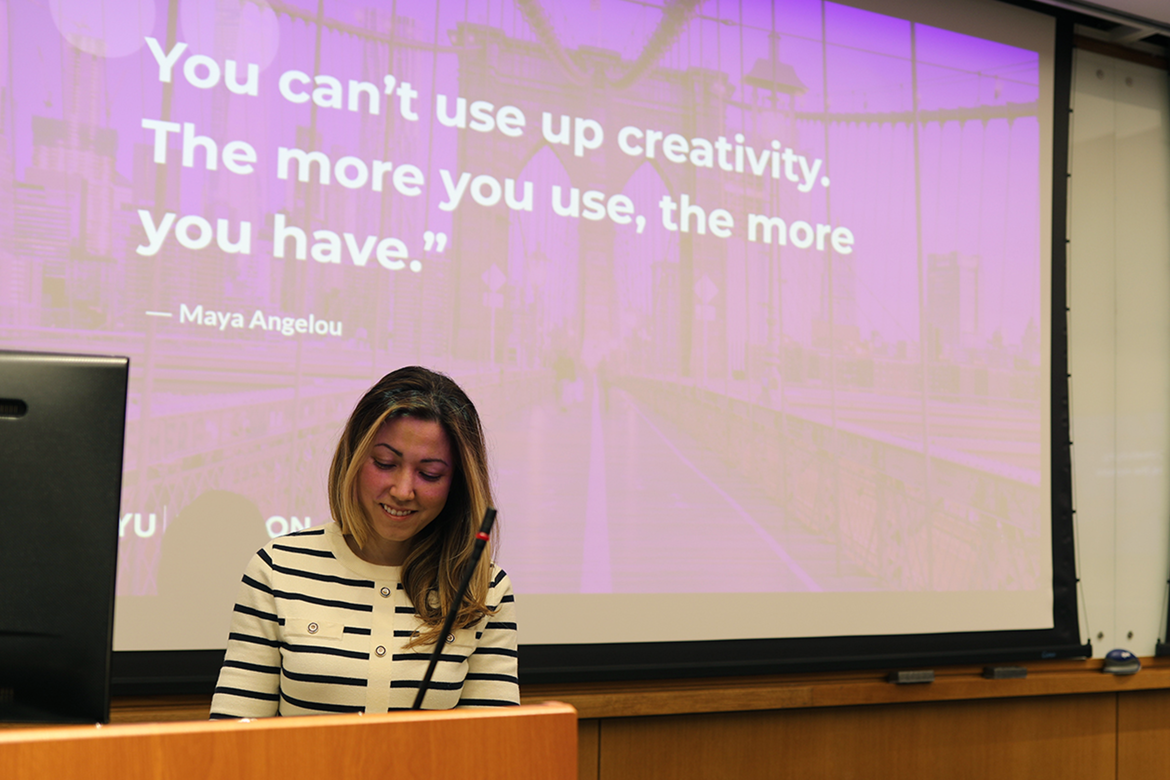 Faculty member Piper Henriques stands at a podium in front of a large screen displaying a Maya Angelou quote reading : "You can't use up creativity. The more you use, the more you have.", and a bridge image in the background.
