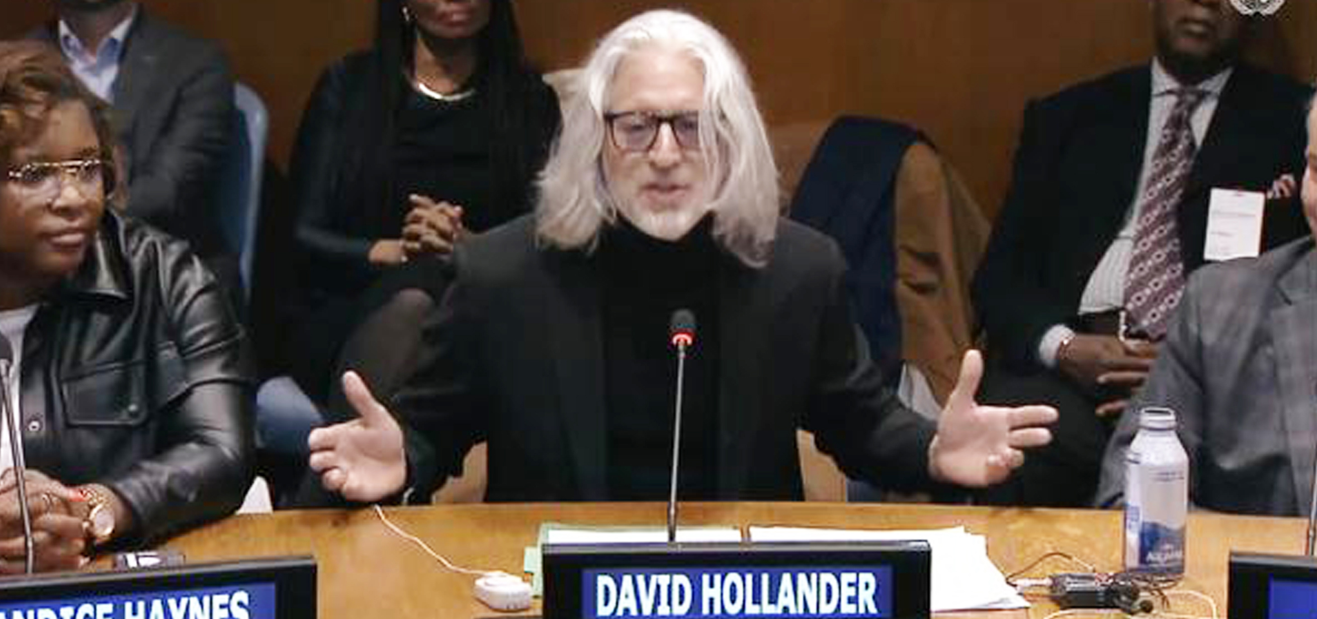 Addressing the U.N. at first every World Basketball Day, David Hollander speaks into a microphone with arms open while sat at a table with his name tag in front of him