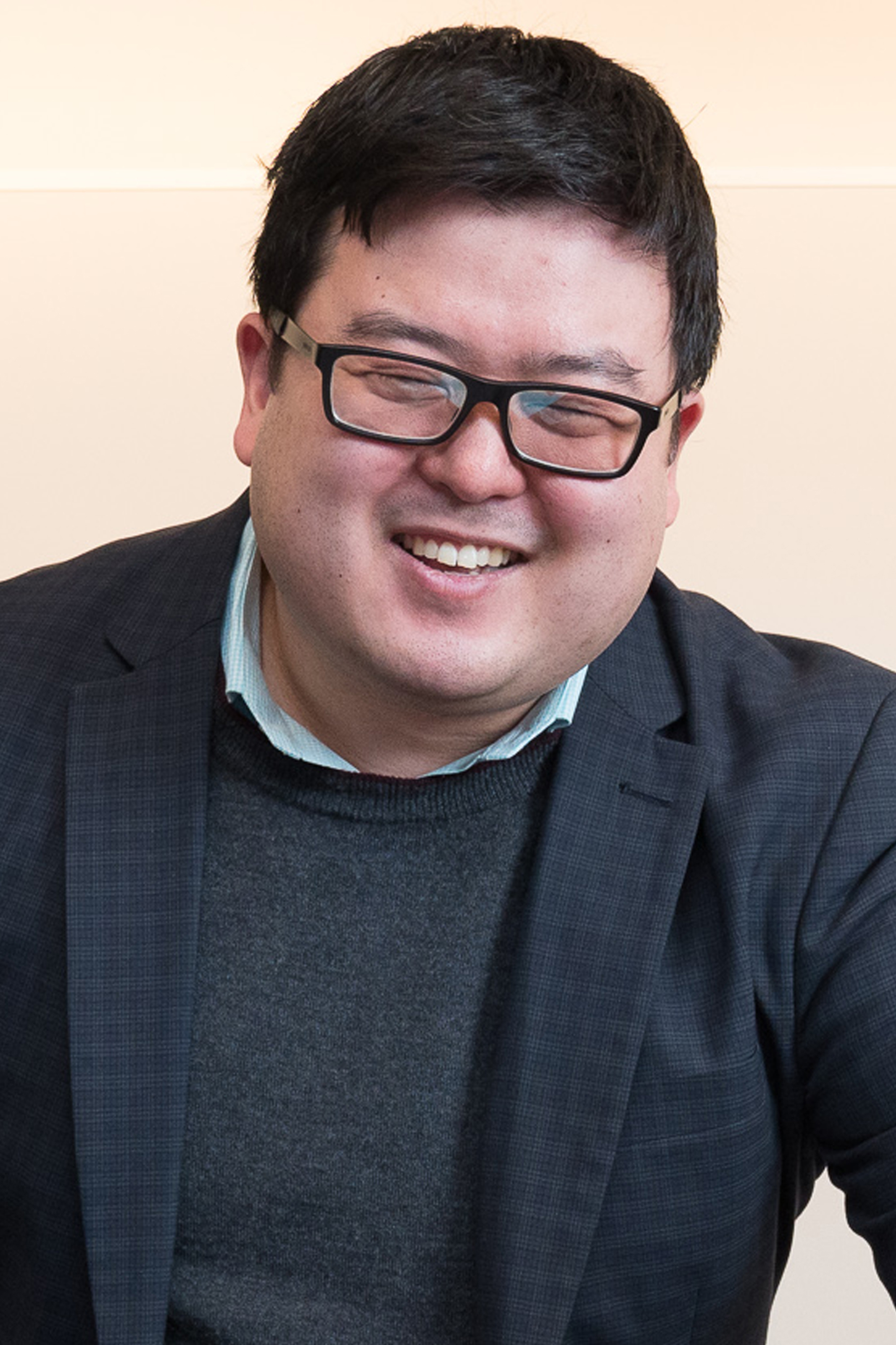 Jason Chung smiles while wearing a dark grey blazer and grey sweater for a headshot in front of a solid white background