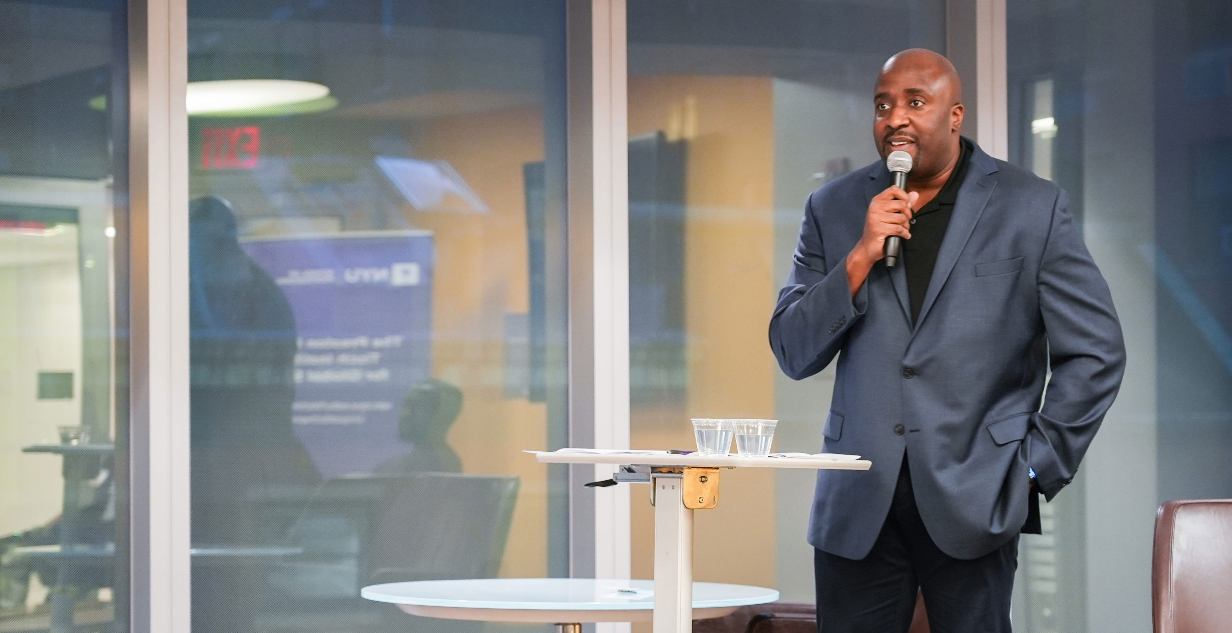 Man in a blue suit speaks confidently into a microphone indoors. He stands beside a high table with water glasses, conveying a professional and engaging tone.