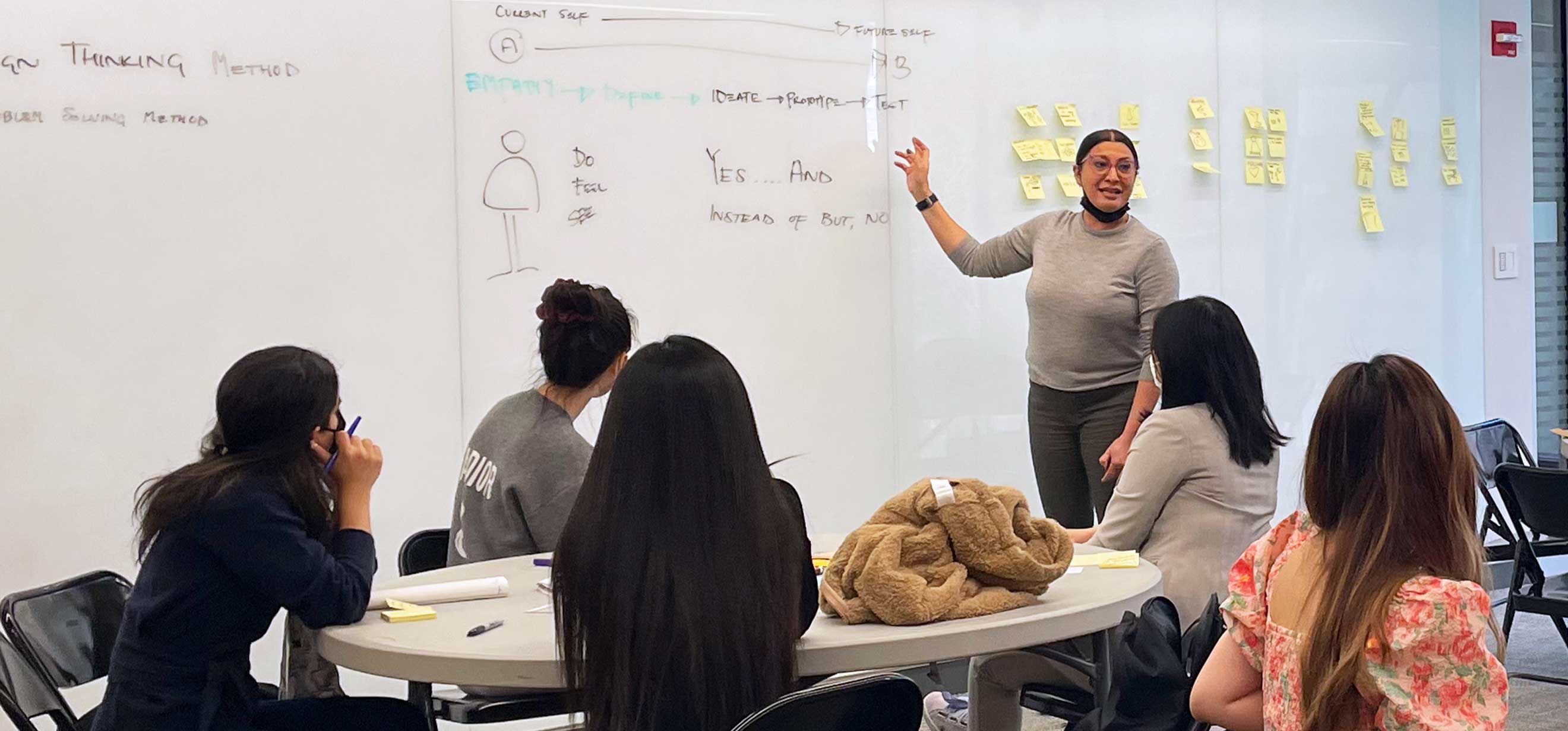 A woman stands at a whiteboard covered with sticky notes, talking to a group of seated students.