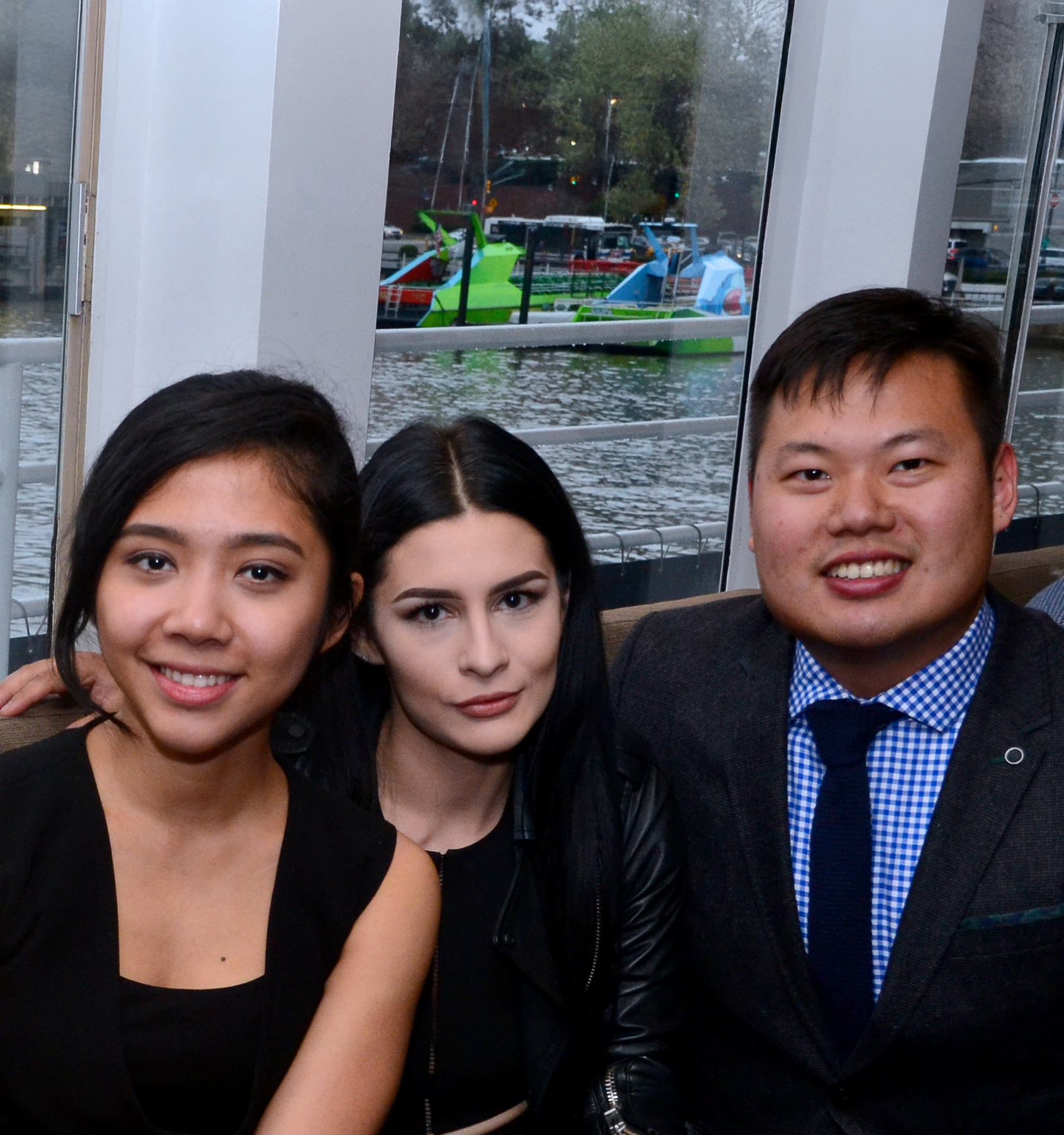 Three NYU SPS external scholarship students pose for a photo in front of a window looking out to a body of water with watercraft in the background